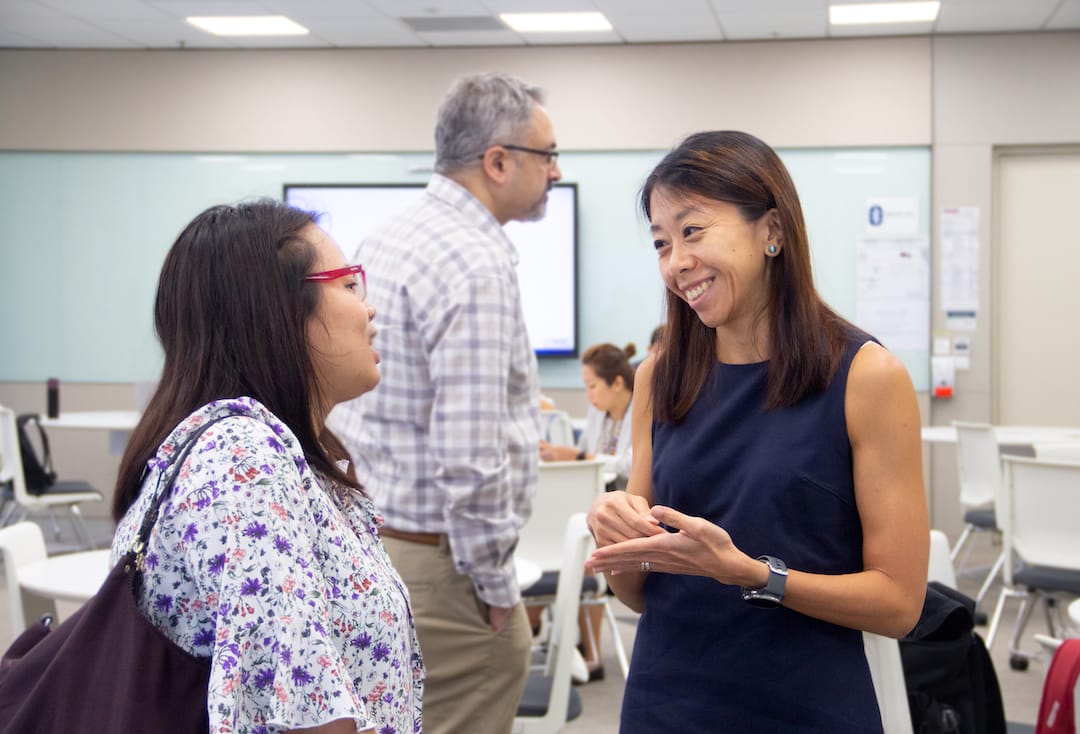 Two women stand conversing in an office, one in a floral shirt and the other in a sleeveless navy dress.